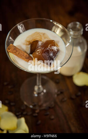 weiße Cocktail übergossen Zimtstange und Kaffee-Eiswürfel in ein Martini-Glas auf eine hölzerne Kulisse umgeben von Rosenblüten Stockfoto