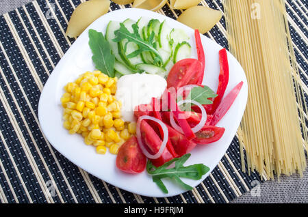 Weißen Teller mit frischen Tomaten, Gurkenscheiben, Mais, Parmeggiano und Zwiebeln neben Zutaten für Spaghetti auf einem gestreiften Hintergrund Stockfoto