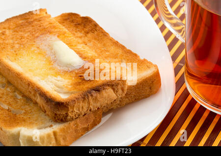 Warmen Toast mit Butter auf einem weißen Teller und eine transparente Tasse Tee Nahaufnahme Stockfoto