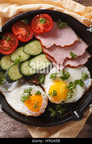 Warmes Frühstück: Spiegelei mit Schinken und frischem Gemüse in einer Pfanne auf dem Tisch. Vertikale Ansicht von oben Stockfoto