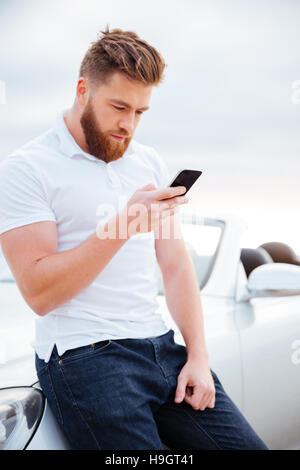 Konzentrierte bärtigen Jüngling mit Smartphone stützte sich auf seinem Auto Stockfoto