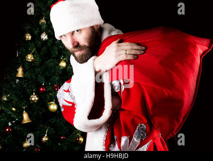 schlechte brutale Weihnachtsmann trägt eine Tasche und gehässig, lächelnd auf dem Hintergrund der Weihnachtsbaum Stockfoto