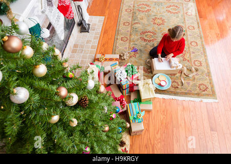 Kleines Mädchen sitzt auf einem Teppich zu Hause unter einem Weihnachtsbaum und Haufen von bunte Geschenke mit ihren speziellen Wunsch neben an den Weihnachtsmann schreiben Stockfoto