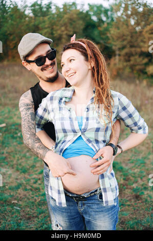 junge Kerl in eine Kappe solar Oskah mit Bart Schnurrbart und tätowiert, seine schwangere Mädchen in Dreadlocks, im Freien im Herbst in die Kamera posiert, Stockfoto
