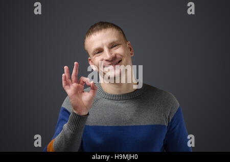 Schöner junger Mann zeigt Geste 'OK'. Emotionale Fotos im Studio auf einem schwarzen Hintergrund. Stockfoto