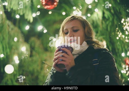 Glückliche junge Dame mit Tasse Kaffee gegen beleuchtete Weihnachtsbaum, geringe Schärfentiefe Stockfoto