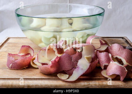 Rote Kartoffeln schält und geschälte Kartoffeln im Glas Schüssel mit Wasser auf Holzbrett von Seite Stockfoto