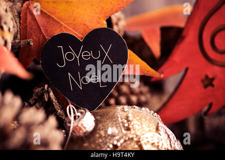 der Text Joyeux Noel, Frohe Weihnachten, geschrieben Französisch in einem herzförmigen Schild und einige Tannenzapfen und Weihnachten Kugeln Stockfoto
