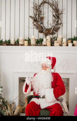Glücklich Weihnachtsmann eine Nachricht auf Ihrem Handy eingeben und lächelnd beim Sitzen auf seinem Stuhl mit Kamin. Weihnachtsbaum im Hintergrund Stockfoto