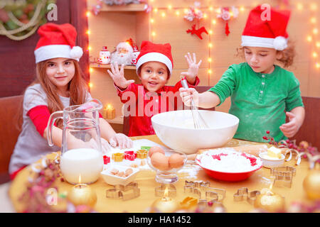 Glückliche Kinder Weihnachtskekse zu Hause machen, niedliche kleine Kinder tragen rote Weihnachtsmann Mützen, festliche Süßigkeiten, traditionelle Feier vorbereiten Stockfoto