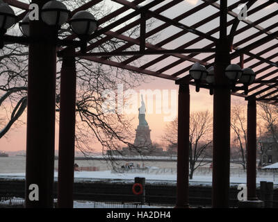 Die legendäre Freiheitsstatue steht bei Sonnenuntergang mit Blick auf Ellis Island, das Symbol für Freiheit und Demokratie in den Vereinigten Staaten. Dieses Wahrzeichen ist seit langem ein willkommenes Symbol für Einwanderer, die nach New York City kommen, und markiert einen historischen Einstiegspunkt nach Amerika. Stockfoto