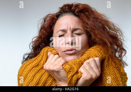 Kalten junge Frau in eine warme gestrickte wollene Winter Top mit schmerzverzerrtem Gesicht und ihre Augen geschlossen, als sie versucht, Aufwärmen, kuscheln hautnah auf weiß Stockfoto