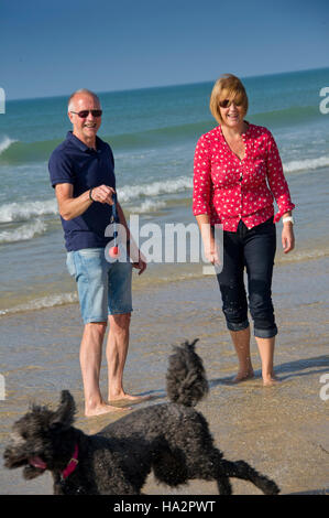 Ein reifes Paar (männlich & weiblich) das Tragen von Kleidung mit ihrem Hund Spaß an einem sonnigen Strand von Cornwall. Stockfoto