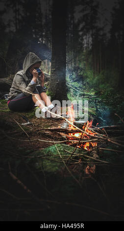 junge Frau trinken heißen Tee durch das Feuer im Wald Stockfoto