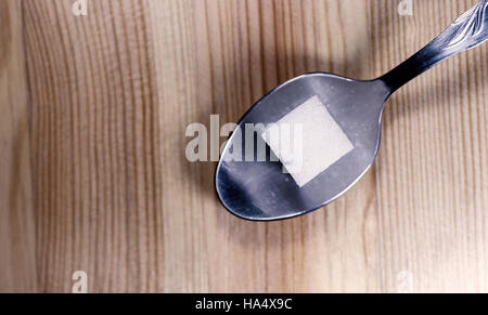 Einzelnen Zucker Cube Klumpen ruht auf einen silbernen Metall Teelöffel auf einem strukturierten Holzküche Desktop. Overhead horizontales Bild. Stockfoto