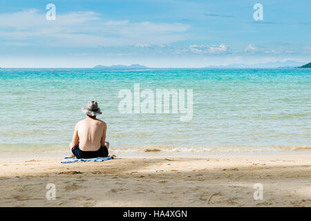 Asiatischer Mann am Strand Rückansicht Kerl Sommer Hut sitzt Blick Meer blauer Himmel, Sommer Urlaub Reisekonzept Freiheit Reisen Strand Meer. Stockfoto