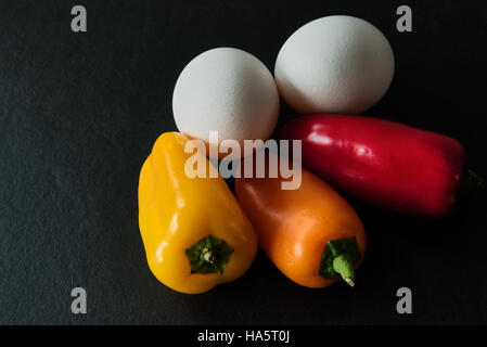 Low-Carb - zwei weißen Eiern mit drei bunten Mini-Paprika auf dunklem Schiefer Hintergrund Stockfoto