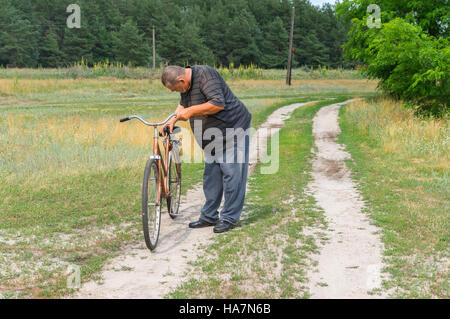 Senior-ukrainische Bauern stehen auf einer Landstraße und Inspektion altes rostigen Fahrrad für immer bereit zur Teilnahme an Grand Tour in dieser Saison Stockfoto