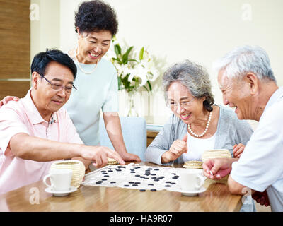 zwei hochrangige asiatische Männer spielen Weiqi (oder Go-Spiel) zu Hause mit ihren Frauen beobachten. Stockfoto