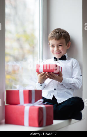 Glücklich süße kleine Junge, tragen, weißes Hemd und Fliege in der Nähe der Fenster holding Geschenk Stockfoto