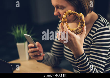 Frau Sesam Bagel zu essen und mit Handy im Büro während der Überstunden Stockfoto