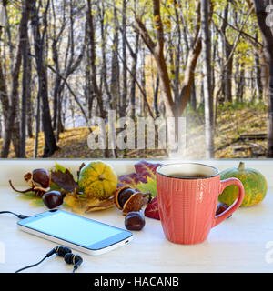 Tasse Kaffee, Smartphone, Kopfhörer und Herbst Blätter auf der Fensterbank. Im Hintergrundfenster mit Herbstlandschaft Stockfoto