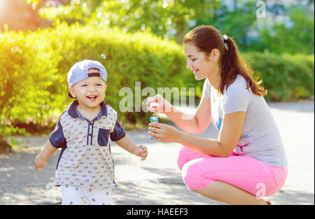 Kleiner Junge mit Mütze im freien laufen. Spaziergang in der Natur mit meiner Mutter und Seifenblasen mit Sonnenlicht. Stockfoto