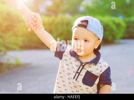 Porträt von ein wenig überrascht Kind in Kappe gehen mit ihrer Mutter auf dem Arm unter freiem Himmel. Spaziergang in der Natur mit Sonnenlicht. Stockfoto