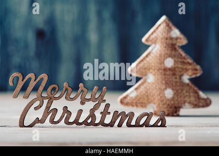 der Text Frohe Weihnachten aus Holz und ein gemütliches Ornament in Form eines Weihnachtsbaumes auf einer rustikalen Holzoberfläche Stockfoto