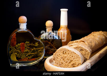 Zusammensetzung auf dunklem Hintergrund. Holzplatte mit frisch geschnittenem Brot und kleine Glasflaschen mit Öl und Gewürzen. Stockfoto