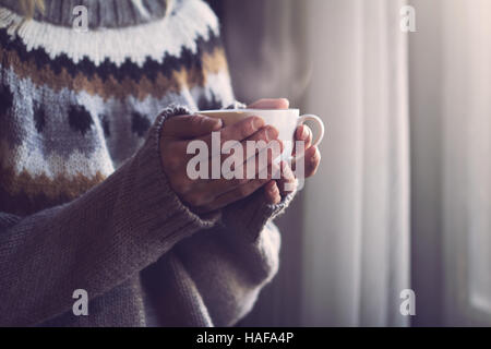 Frau im grauen Strickpullover mit traditionellen Motiven in Händen hält, eine weiße Tasse heißen Kaffee mit Dampf kommt im Licht morgens Stockfoto