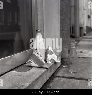 1965, sitzen historische, eine Flasche Bier Größe Glas Milch und zwei Milchtüten auf eine Haustür, mit einem leeren Glas Milch Flasche stehen auf dem Bürgersteig. Stockfoto