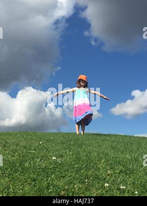 Ein junges Mädchen, das auf dem Grasberg steht, trägt Sommerkleid und Hut mit ausgestreckten Armen Stockfoto