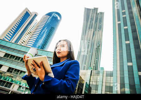 Ernsthafte asiatische junge Geschäftsfrau mit einer Einweg-Kaffeetasse, Kaffee zu trinken, und Tablet gegen städtische Stadt Wolkenkratzer in ihren Händen hält Stockfoto