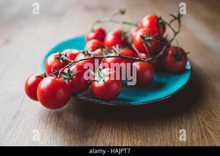 Cherry-Tomaten am Rebstock. Blaue Teller mit Cherry-Tomaten auf Holztisch. Stockfoto