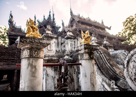 Shwenandaw Kloster Goldene Figuren Mandalay Myanmar // MANDALAY, Myanmar — Goldene Figuren aus der buddhistischen Ikonographie sitzen auf kleinen Sockeln, die eine Glocke im Innenhof des Shwenandaw Klosters (auch bekannt als Shwe Nandaw Kyaung) stützen. Das Kloster zeigt komplexe Teakholzschnitzereien, die buddhistische Mythologie und Jataka-Geschichten in seiner gesamten Struktur darstellen. Das 1878 erbaute Kloster war ursprünglich König Mindon Mindons private königliche Wohnung im Mandalay Palace Komplex, bevor es nach dem Tod des Königs in ein buddhistisches Kloster umgewandelt wurde. Die Struktur repräsentiert die einzige überlebende Stockfoto