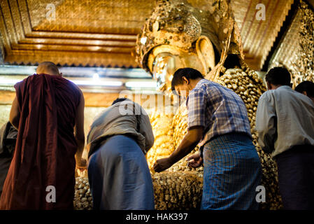 Mahamuni Buddha Bild Gold Blatt Anwendung Mandalay Myanmar // MANDALAY, Myanmar — Männer wenden Blattgold auf das Mahamuni Buddha Bild im inneren Heiligtum des Mahamuni Buddha Tempels an (auch bekannt als Mahamuni Pagoda). Die Bronzestatue mit einem Gewicht von 6,5 Tonnen und einer Höhe von 3,82 Metern gilt als eine von nur fünf originalen Gestalten Buddhas, die zu seinen Lebzeiten entstanden sind. Im Laufe der Jahrhunderte haben Gläubige etwa 15 Zentimeter Blattgold auf das Bild aufgetragen, wobei nur männliche Anbeter die Statue direkt berühren und vergolden dürfen. Der 1785 von König Bodawpaya erbaute Tempel beherbergt das Stockfoto