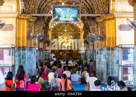 Mahamuni Buddha Tempel Goldblatt Zeremonie Mandalay Myanmar // MANDALAY, Myanmar – Gläubige und Pilger treffen sich in der äußeren Halle des Mahamuni Buddha Tempels (auch bekannt als Mahamuni Pagode), um den Prozess der Anwendung von Blattgold auf dem verehrten Mahamuni Buddha Bild zu beobachten. Der Tempel hat traditionelle Geschlechterbeschränkungen, wonach nur Männer das innere Heiligtum betreten und direkt an der Anwendung von Blattgold auf die Bronzestatue teilnehmen dürfen, die 6,5 Tonnen wiegt und mit etwa 15 Zentimeter Blattgold aus Jahrhunderten von Andachtsopfern bedeckt ist. Frauen und andere Stockfoto