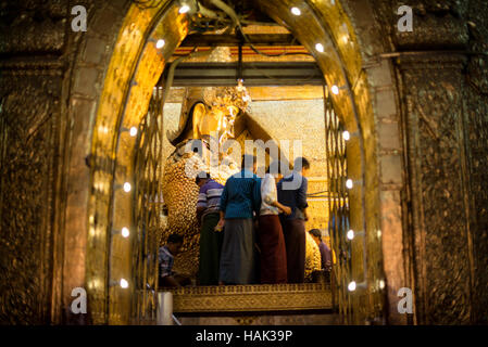 Mahamuni Buddha Tempel Gold Blatt Anwendung Mandalay Myanmar // MANDALAY, Myanmar — Männer wenden Blattgold auf das Mahamuni Buddha Bild im inneren Heiligtum des Mahamuni Buddha Tempels an (auch bekannt als Mahamuni Pagoda). Die Bronzestatue mit einem Gewicht von 6,5 Tonnen und einer Höhe von 3,82 Metern gilt als eine von nur fünf originalen Gestalten Buddhas, die zu seinen Lebzeiten entstanden sind. Im Laufe der Jahrhunderte haben Gläubige etwa 15 Zentimeter Blattgold auf das Bild aufgetragen, wobei nur männliche Anbeter die Statue direkt berühren und vergolden dürfen. Der 1785 von König Bodawpaya erbaute Tempel beherbergt diesen Stockfoto