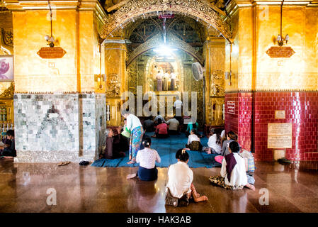 Mahamuni Buddha Tempel Gold Blatt Anwendung Mandalay Myanmar // MANDALAY, Myanmar — Gläubige und Pilger treffen sich in der äußeren Halle des Mahamuni Buddha Tempels (auch bekannt als Mahamuni Pagoda), um den Prozess der Anwendung von Blattgold auf dem verehrten Mahamuni Buddha Bild zu beobachten. Der Tempel hat traditionelle Geschlechterbeschränkungen, wonach nur Männer das innere Heiligtum betreten und direkt an der Anwendung von Blattgold auf die Bronzestatue teilnehmen dürfen, die 6,5 Tonnen wiegt und mit etwa 15 Zentimeter Blattgold aus Jahrhunderten von Andachtsopfern bedeckt ist. Frauen und andere Stockfoto