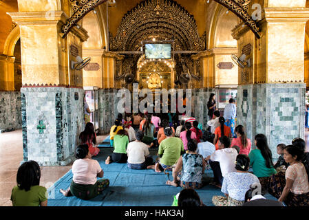 Mahamuni Buddha Tempel Pilger Mandalay Myanmar // MANDALAY, Myanmar — Gläubige und Pilger treffen sich in der äußeren Halle des Mahamuni Buddha Tempels (auch bekannt als Mahamuni Pagode), um den Prozess der Anwendung von Blattgold auf dem verehrten Mahamuni Buddha Bild zu beobachten. Der Tempel hat traditionelle Geschlechterbeschränkungen, wonach nur Männer das innere Heiligtum betreten und direkt an der Anwendung von Blattgold auf die Bronzestatue teilnehmen dürfen, die 6,5 Tonnen wiegt und mit etwa 15 Zentimeter Blattgold aus Jahrhunderten von Andachtsopfern bedeckt ist. Frauen und andere Besucher o Stockfoto
