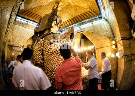 Mahamuni Buddha Bild Gold Blatt Anwendung Mandalay Myanmar // MANDALAY, Myanmar — Männer wenden Blattgold auf das Mahamuni Buddha Bild im inneren Heiligtum des Mahamuni Buddha Tempels an (auch bekannt als Mahamuni Pagoda). Die Bronzestatue mit einem Gewicht von 6,5 Tonnen und einer Höhe von 3,82 Metern gilt als eine von nur fünf originalen Gestalten Buddhas, die zu seinen Lebzeiten entstanden sind. Im Laufe der Jahrhunderte haben Gläubige etwa 15 Zentimeter Blattgold auf das Bild aufgetragen, wobei nur männliche Anbeter die Statue direkt berühren und vergolden dürfen. Der 1785 von König Bodawpaya erbaute Tempel beherbergt das Stockfoto