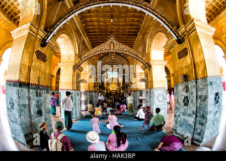 Mahamuni Buddha Tempel Pilger Mandalay Myanmar // MANDALAY, Myanmar — Gläubige und Pilger treffen sich in der äußeren Halle des Mahamuni Buddha Tempels (auch bekannt als Mahamuni Pagode), um den Prozess der Anwendung von Blattgold auf dem verehrten Mahamuni Buddha Bild zu beobachten. Der Tempel hat traditionelle Geschlechterbeschränkungen, wonach nur Männer das innere Heiligtum betreten und direkt an der Anwendung von Blattgold auf die Bronzestatue teilnehmen dürfen, die 6,5 Tonnen wiegt und mit etwa 15 Zentimeter Blattgold aus Jahrhunderten von Andachtsopfern bedeckt ist. Frauen und andere Besucher o Stockfoto