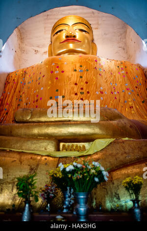 BAGAN, Myanmar — Eine kolossale Buddha-Statue, Die in Safrangewänder gehüllt ist, befindet sich innerhalb der engen Grenzen des Manuha-Tempels, eines von drei monumentalen Buddha-Bildern, die in den östlichen Kammern des Tempels untergebracht sind. Der Tempel wurde 1067 n. Chr. von dem Gefangenen Mon König Manuha nach seiner Niederlage durch König Anawrahta aus dem heidnischen Königreich erbaut und verfügt über bewusst beengte Räume, die die psychische Not des inhaftierten Königs symbolisieren. Der zentral sitzende Buddha ist 46 Fuß (14 Meter) hoch und stellt den Buddha im Posten „die Erde zum Zeugen rufen“ dar Stockfoto
