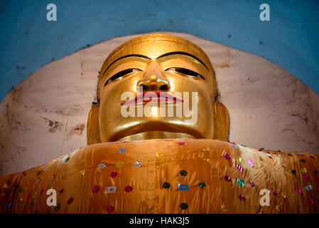 BAGAN, Myanmar — Eine kolossale Buddha-Statue, Die in Safrangewänder gehüllt ist, befindet sich innerhalb der engen Grenzen des Manuha-Tempels, eines von drei monumentalen Buddha-Bildern, die in den östlichen Kammern des Tempels untergebracht sind. Der Tempel wurde 1067 n. Chr. von dem Gefangenen Mon König Manuha nach seiner Niederlage durch König Anawrahta aus dem heidnischen Königreich erbaut und verfügt über bewusst beengte Räume, die die psychische Not des inhaftierten Königs symbolisieren. Der zentral sitzende Buddha ist 46 Fuß (14 Meter) hoch und stellt den Buddha im Posten „die Erde zum Zeugen rufen“ dar Stockfoto