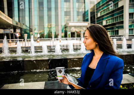 Ernsthafte asiatische junge Geschäftsfrau mit einer Einweg-Kaffeetasse, Kaffee zu trinken, und Tablet gegen städtische Stadt Wolkenkratzer in ihren Händen hält Stockfoto