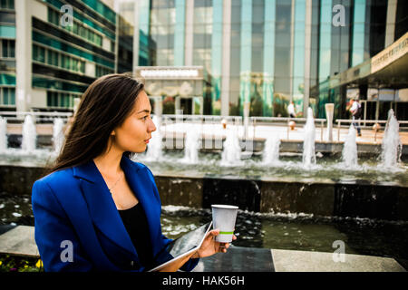 Ernsthafte asiatische junge Geschäftsfrau mit einer Einweg-Kaffeetasse, Kaffee zu trinken, und Tablet gegen städtische Stadt Wolkenkratzer in ihren Händen hält Stockfoto