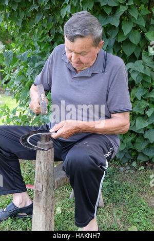 Bauer mit Hammer und Eisen-Tool auf dem Baumstumpf ist seine Sense in Nedelisce, Kroatien schärfen. Stockfoto