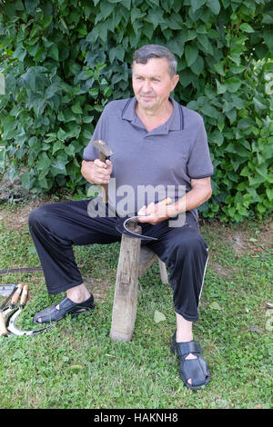 Bauer mit Hammer und Eisen-Tool auf dem Baumstumpf ist seine Sense in Nedelisce, Kroatien schärfen. Stockfoto