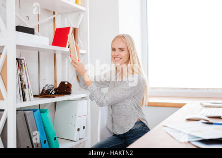 Lächelnde junge Frau nehmen Buch aus einem Regal im Büro Stockfoto